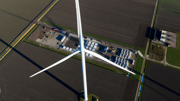 Aerial view of a large white wind turbine standing next to a battery energy storage system facility surrounded by vast agricultural fields in Dronten Flevoland Netherlands.