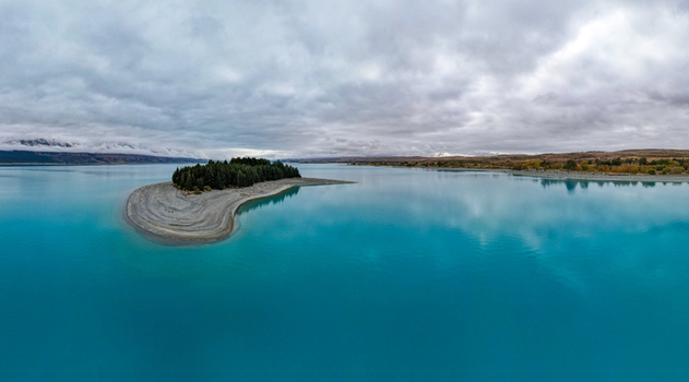 Aerial view of the turquoise waters of Lake Pukaki with a small island covered in pine trees surrounded by distant mountains under a cloudy sky in Twizel Canterbury Region New Zealand.