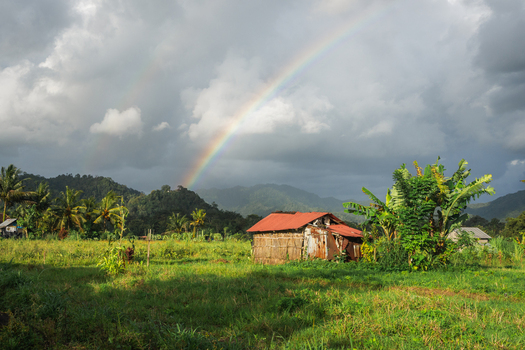 View of a rustic wooden hut with a red roof in a lush green field under a vibrant rainbow and dramatic cloudy sky Sidemen Bali Indonesia. View of a rustic wooden hut with a red roof in a lush green field under a vibrant rainbow and dramatic cloudy sky Sidemen Bali Indonesia.