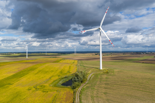 Aerial view of wind turbines standing in vast agricultural fields with a small pond and a dirt path under a dramatic cloudy sky in Pomeranian Voivodeship Poland.