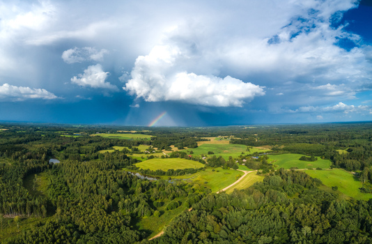 Aerial view of a lush green landscape featuring dense forests rolling meadows and a small rainbow appearing under dramatic storm clouds in Latvia. Aerial view of a lush green landscape featuring dense forests rolling meadows and a small rainbow appearing under dramatic storm clouds in Latvia.