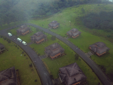 Aerial view of traditional style bungalows with dark textured roofs nestled on verdant hills connected by winding asphalt roads under a misty sky Obudu Cross River Nigeria.