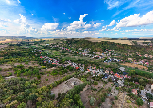 Aerial view of Csabdi village in Hungary with traditional homes in a wooded valley beneath rolling hills patchwork farmland and a bright early autumn sky.