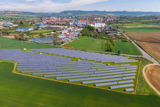 Aerial view of a solar farm with rows of photovoltaic panels in green fields near industrial buildings and rolling hills in Kyjov South Moravian Region Czechia.
