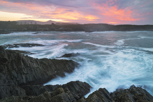 View of the rugged coastline near Illa Pancha with waves crashing against rocks under a pink and orange sunset sky Lugo Lugo Spain.