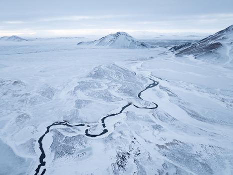 Aerial view of a winding river cutting through a vast snow covered landscape with distant mountains under a soft winter sky in Egilsstadir Mulathing Iceland. Aerial view of a winding river cutting through a vast snow covered landscape with distant mountains under a soft winter sky in Egilsstadir Mulathing Iceland.