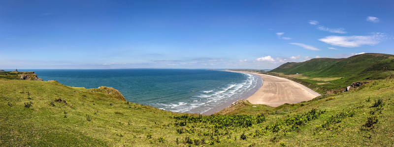 Aerial view of Rhossili Beach with its long sandy shoreline rolling green hills and turquoise sea under a clear blue sky Rhossili United Kingdom. Aerial view of Rhossili Beach with its long sandy shoreline rolling green hills and turquoise sea under a clear blue sky Rhossili United Kingdom.