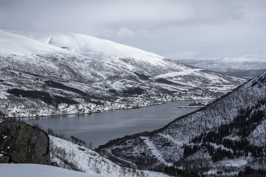 View of the snow covered mountains surrounding the calm waters of a fjord with a small coastal village under an overcast winter sky in Ersfjord Troms Norway. View of the snow covered mountains surrounding the calm waters of a fjord with a small coastal village under an overcast winter sky in Ersfjord Troms Norway.
