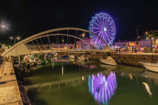 View of a modern white arch bridge over a canal at night with a glowing purple ferris wheel in the background and reflections on the water in Fano Marche Italy. View of a modern white arch bridge over a canal at night with a glowing purple ferris wheel in the background and reflections on the water in Fano Marche Italy.