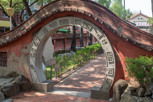 View of the traditional stone moon gate at Chihkan Tower with intricate carvings set in a red wall leading to a garden path in Chihkan Village Tainan City Taiwan. View of the traditional stone moon gate at Chihkan Tower with intricate carvings set in a red wall leading to a garden path in Chihkan Village Tainan City Taiwan.