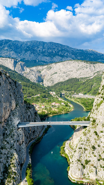 Aerial view of the Cetina River canyon bridge spanning steep limestone cliffs and lush green valley under a blue sky with white clouds in Omis Split Dalmatia County Croatia. Aerial view of the Cetina River canyon bridge spanning steep limestone cliffs and lush green valley under a blue sky with white clouds in Omis Split Dalmatia County Croatia.