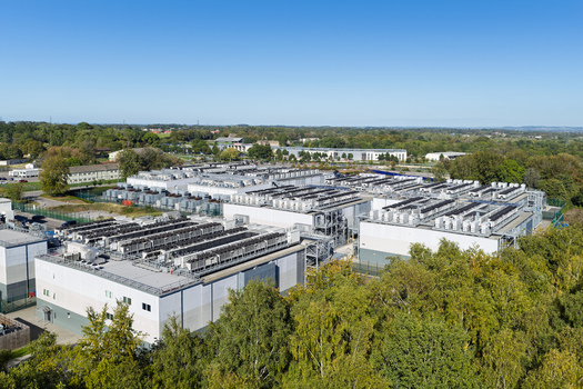 Aerial view of a large data center complex with multiple buildings and rooftop cooling units surrounded by lush forest in the United Kingdom.