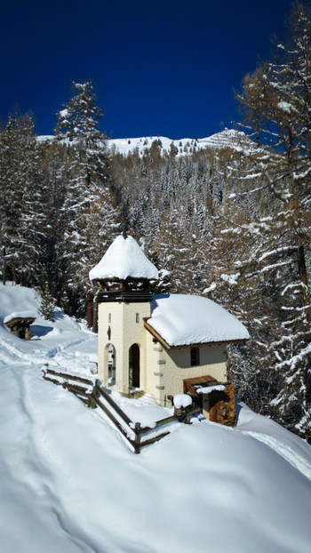 Aerial view of a snow laden chapel standing serene against a backdrop of snow covered mountains and trees Grimentz Valais Switzerland.