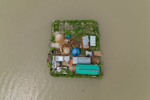 Mithamain Upazila Bangladesh 09 May 2026 Aerial view of a small isolated homestead island with corrugated metal roofs and haystacks surrounded by floodwaters.