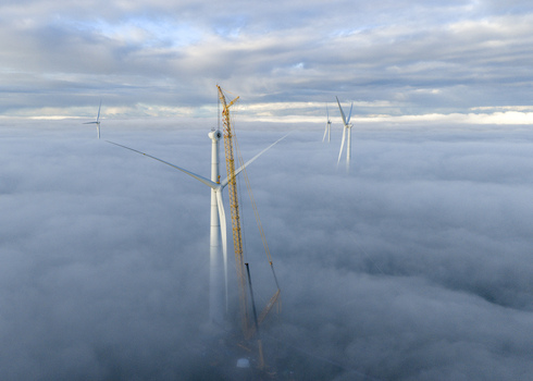 Aerial view of wind turbines and a tall yellow crane rising above a dense sea of clouds under a cloudy sky.