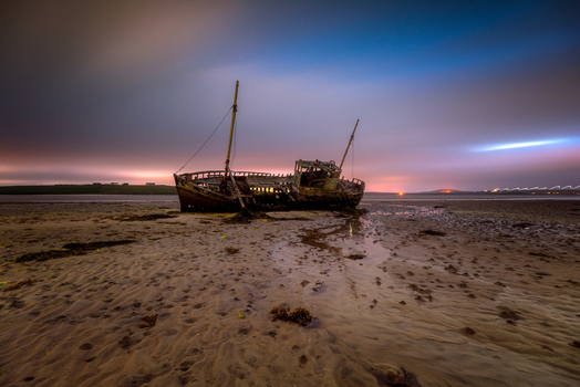 View of the historic Rossport shipwreck resting on the sandy beach under a twilight sky with purple and blue hues in Ross Port County Mayo Ireland. View of the historic Rossport shipwreck resting on the sandy beach under a twilight sky with purple and blue hues in Ross Port County Mayo Ireland.