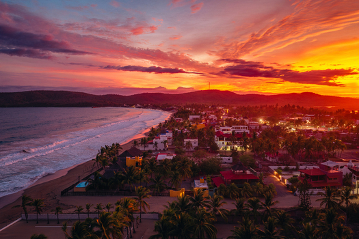 Aerial view of Playa Punta Perula at sunset with vibrant orange and pink clouds over the coastline palm trees and buildings in Perula Jalisco Mexico. Aerial view of Playa Punta Perula at sunset with vibrant orange and pink clouds over the coastline palm trees and buildings in Perula Jalisco Mexico.