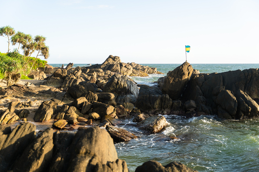 View of rugged rock formations along the coastline with ocean waves crashing against the shore and a small flag under a clear sky in Galle Southern Province Sri Lanka.