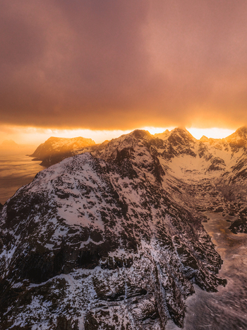Aerial view of jagged snow dusted peaks pierce the horizon as a fiery sunset paints the sky in hues of gold and crimson Nordland Norway.
