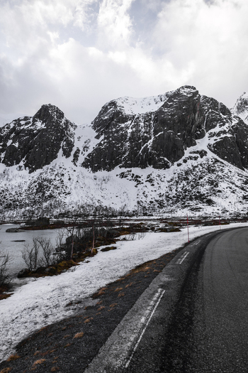 View of a winding asphalt road leading towards snow covered rugged mountains and a calm lake under a cloudy sky in Kvaloya Troms Norway. View of a winding asphalt road leading towards snow covered rugged mountains and a calm lake under a cloudy sky in Kvaloya Troms Norway.