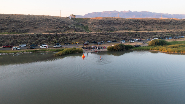 Aerial view of a person paddleboarding on a calm lake near a shoreline with a crowd of people and parked vehicles set against rolling hills in Cody Wyoming United States.