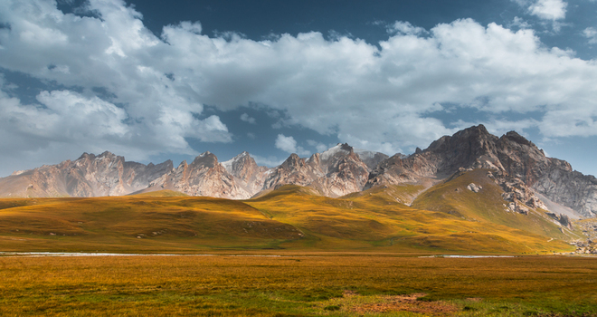 View of the majestic Kol Suu mountain range with golden grassy plains under a dramatic cloudy sky in At Bashi District Naryn Region Kyrgyzstan. View of the majestic Kol Suu mountain range with golden grassy plains under a dramatic cloudy sky in At Bashi District Naryn Region Kyrgyzstan.