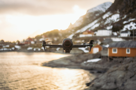 View of a drone hovering above the water as the sun sets behind snowy mountains and orange houses in Tind Nordland Norway.