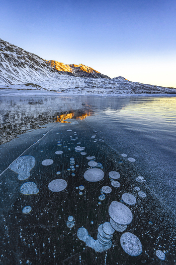 View of frozen lake with methane bubbles trapped under the ice and snow covered mountains during sunset in Madesimo Lombardy Italy. View of frozen lake with methane bubbles trapped under the ice and snow covered mountains during sunset in Madesimo Lombardy Italy.