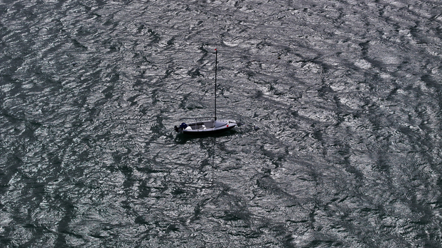 Aerial view of a small white boat with a tall mast floating on the dark rippled surface of the sea in Carantec Brittany France. Aerial view of a small white boat with a tall mast floating on the dark rippled surface of the sea in Carantec Brittany France.