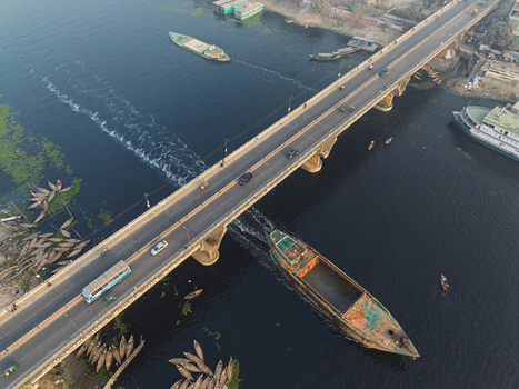 Dhaka Division Bangladesh 23 January 2026 Aerial view of the Buriganga River with a large cargo boat passing under a busy bridge and traditional wooden boats along the banks.