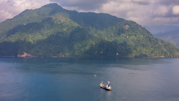 Aerial view of Pantai Karanggongso featuring a cargo ship sailing in the calm blue bay near lush green mountains under a cloudy sky Watulimo East Java Indonesia.