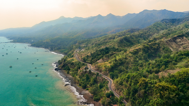 Aerial view of a winding coastal road along the lush green mountainside meeting the turquoise sea in Sukabumi Indonesia. Aerial view of a winding coastal road along the lush green mountainside meeting the turquoise sea in Sukabumi Indonesia.