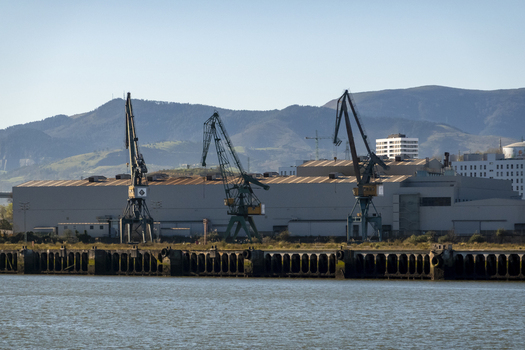 View of an industrial port area featuring large cargo cranes and warehouse buildings against a backdrop of rolling hills in Greater Bilbao Basque Country Spain. View of an industrial port area featuring large cargo cranes and warehouse buildings against a backdrop of rolling hills in Greater Bilbao Basque Country Spain.