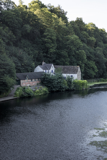 View of a historic stone building nestled along the riverbank surrounded by dense woodland in Durham England United Kingdom. View of a historic stone building nestled along the riverbank surrounded by dense woodland in Durham England United Kingdom.
