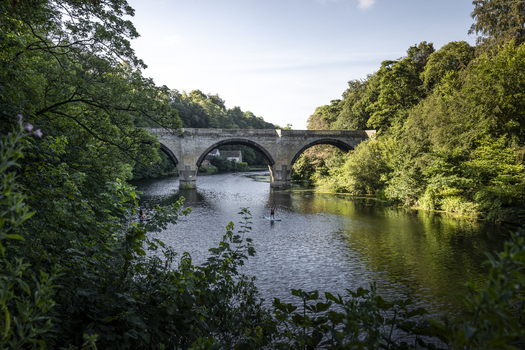 View of Prebends Bridge spanning the River Wear surrounded by lush green trees with a person paddleboarding on the calm water in Durham England United Kingdom. View of Prebends Bridge spanning the River Wear surrounded by lush green trees with a person paddleboarding on the calm water in Durham England United Kingdom.