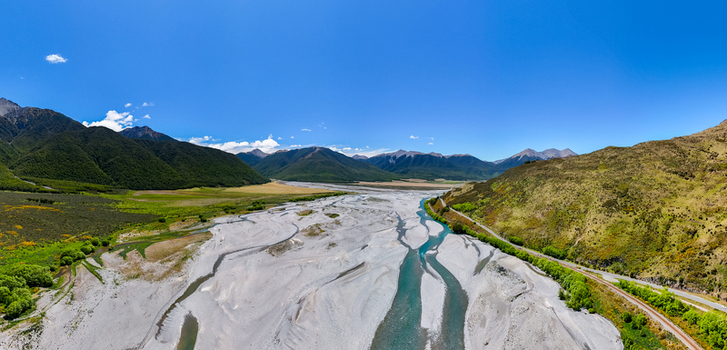 Aerial view of a braided river with turquoise water channels and gravel beds winding through the landscape surrounded by green hills and mountains in Canterbury Region New Zealand.