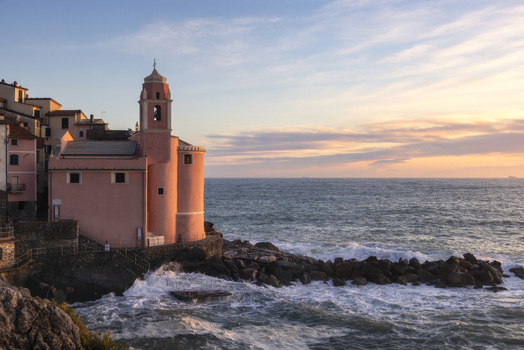 View of the pink Church of San Giorgio perched on the rocky coastline with waves crashing against the shore under a soft sunset sky Tellaro Liguria Italy. View of the pink Church of San Giorgio perched on the rocky coastline with waves crashing against the shore under a soft sunset sky Tellaro Liguria Italy.