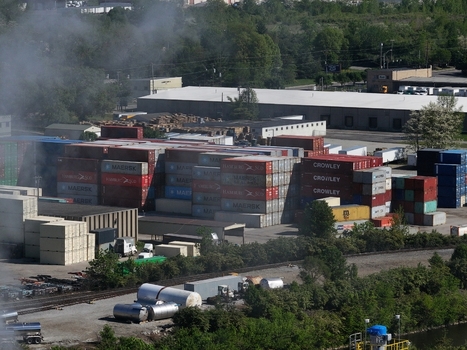 Cincinnati United States 19 April 2026 Aerial view of stacked shipping containers in an industrial yard with smoke rising in the background and lush green trees.