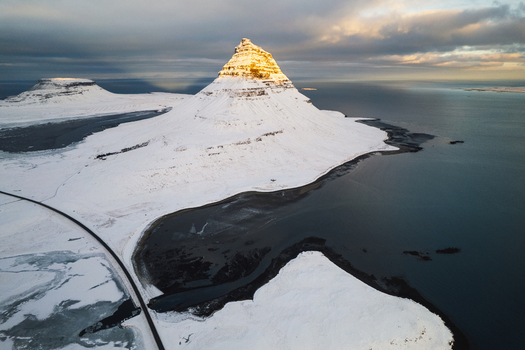 Aerial view of snow dusted Kirkjufell mountain under a glow of soft sunlight contrasting with the dark reflective waters Kirkjufell Grundarfjar&eth;arb&aelig;r Iceland.