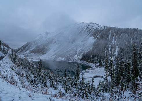 View of a snow covered mountain peak and a calm alpine lake surrounded by dense evergreen forest under a misty overcast sky in United States. View of a snow covered mountain peak and a calm alpine lake surrounded by dense evergreen forest under a misty overcast sky in United States.