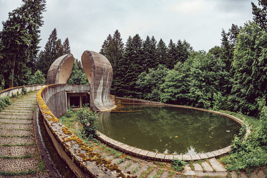 View of the Korcanica Memorial Zone featuring a curved concrete monument and a circular pool surrounded by a dense pine forest at Sanski Most Federation of Bosnia Bosnia and Herzegovina. View of the Korcanica Memorial Zone featuring a curved concrete monument and a circular pool surrounded by a dense pine forest at Sanski Most Federation of Bosnia Bosnia and Herzegovina.
