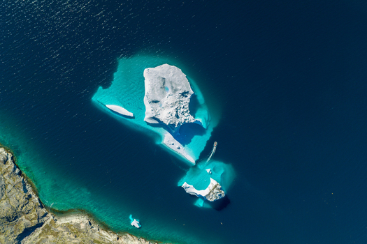 Aerial view of icebergs floating in deep blue arctic waters with a small boat navigating near the ice and a rocky coastline in Greenland. Aerial view of icebergs floating in deep blue arctic waters with a small boat navigating near the ice and a rocky coastline in Greenland.