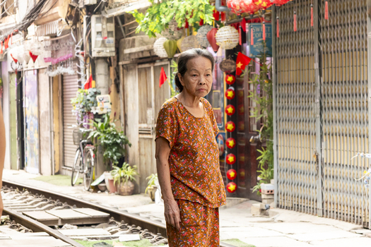 Hanoi Vietnam 11 November 2025 View of an elderly woman standing on the train tracks of Hanoi Train Street with narrow residential buildings hanging lanterns and potted plants.