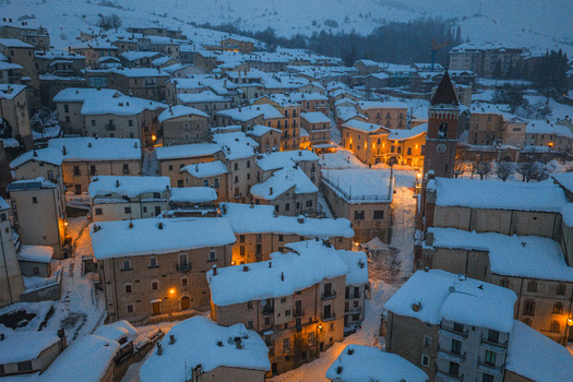 Aerial view of the historic village covered in deep snow with illuminated stone buildings narrow streets and a prominent church tower at dusk in Rivisondoli Abruzzo Italy.