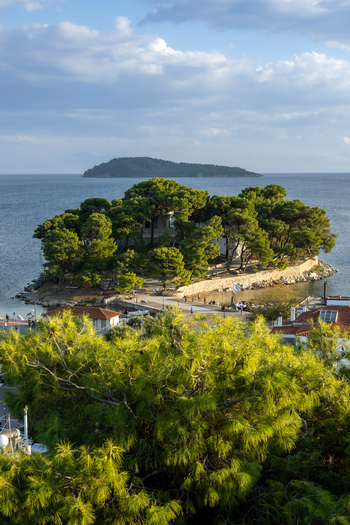 View of a small pine covered island connected by a stone path to the mainland surrounded by the calm blue sea under a clear sky in Lefkada Greece.