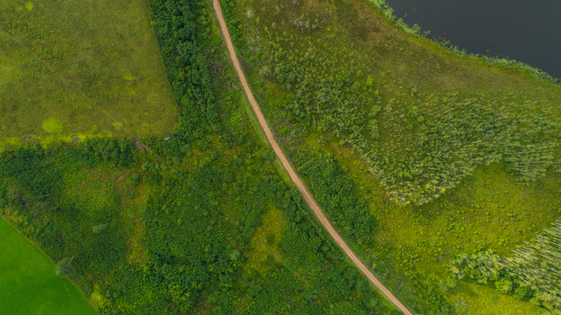 Aerial view of a dirt road cutting through a lush green forest and fields next to a dark lake in Latvia. Aerial view of a dirt road cutting through a lush green forest and fields next to a dark lake in Latvia.