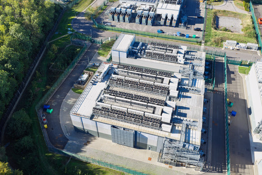 Aerial view of a modern data center facility featuring extensive rooftop cooling units secure perimeter fencing and surrounding green space in the United Kingdom.