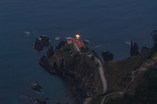 Aerial view of the Cudillero Lighthouse perched on a rugged cliff with its beacon shining over the dark Cantabrian Sea at dusk Cudillero Asturias Spain. Aerial view of the Cudillero Lighthouse perched on a rugged cliff with its beacon shining over the dark Cantabrian Sea at dusk Cudillero Asturias Spain.