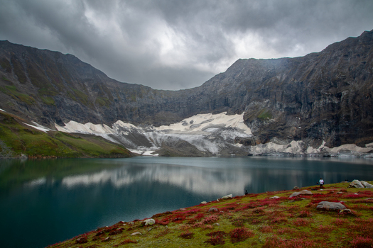 View of Chitta Katha Lake in Neelum Valley surrounded by rugged mountains snow patches and vibrant red alpine vegetation under a cloudy sky in Neelum Kashmir Pakistan. View of Chitta Katha Lake in Neelum Valley surrounded by rugged mountains snow patches and vibrant red alpine vegetation under a cloudy sky in Neelum Kashmir Pakistan.