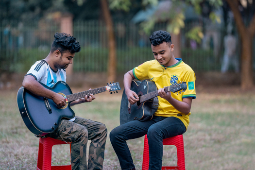 Bogura Rajshahi Division Bangladesh 02 December 2022 View of two young men sitting on red plastic stools in a park playing acoustic guitars while wearing football jerseys.